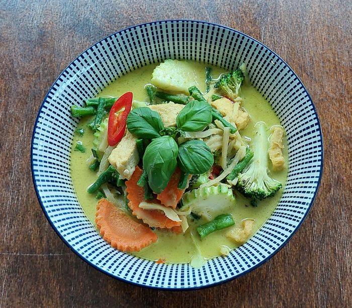 Een kom Thaise groene curry met tofu, broccoli, wortels en sperziebonen, gegarneerd met verse basilicum en een schijfje rode chilipeper.