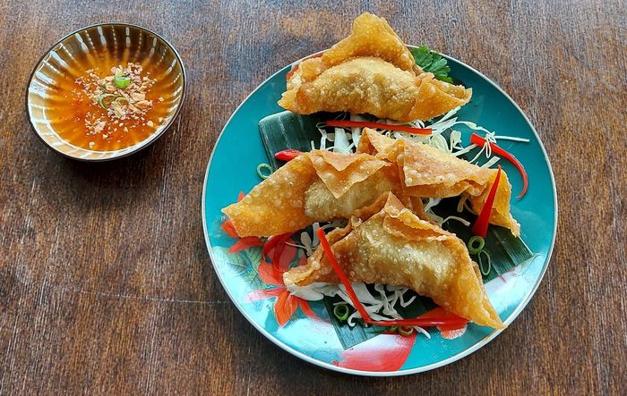 Goudbruin gefrituurde wontons geserveerd op een turquoise bord met een dipsausje, op een donkere, gestructureerde ondergrond.