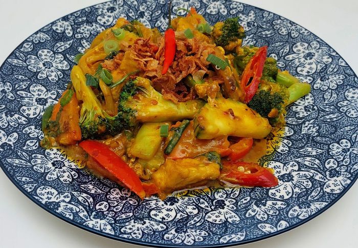 Een kleurrijke roerbak met broccoli, aardappelen en rode chilipepers op een blauw-wit bord.