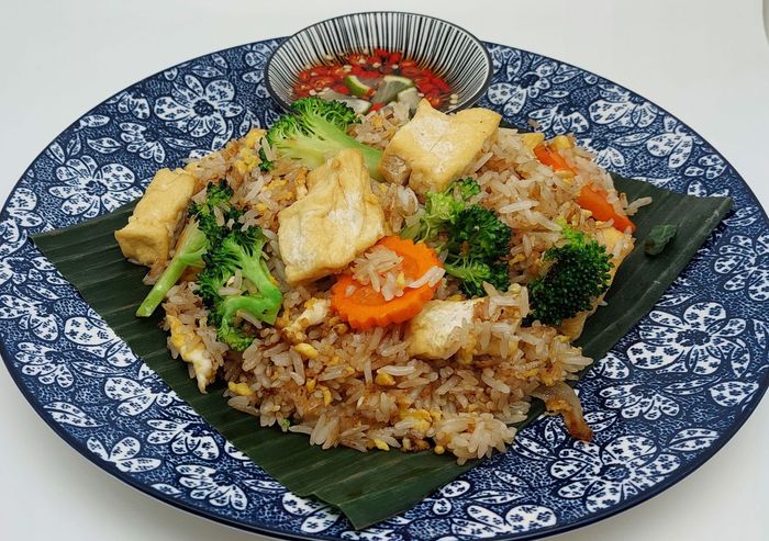 Gebakken rijst met tofu, broccoli en wortelen, geserveerd op een bananenblad op een blauw bord met patroon, met een schaaltje chilisaus erbij.