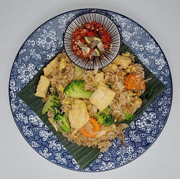 Gebakken rijst met tofu, broccoli en wortelen op een bananenblad, geserveerd met een klein kommetje chili en limoen op een blauw bord.