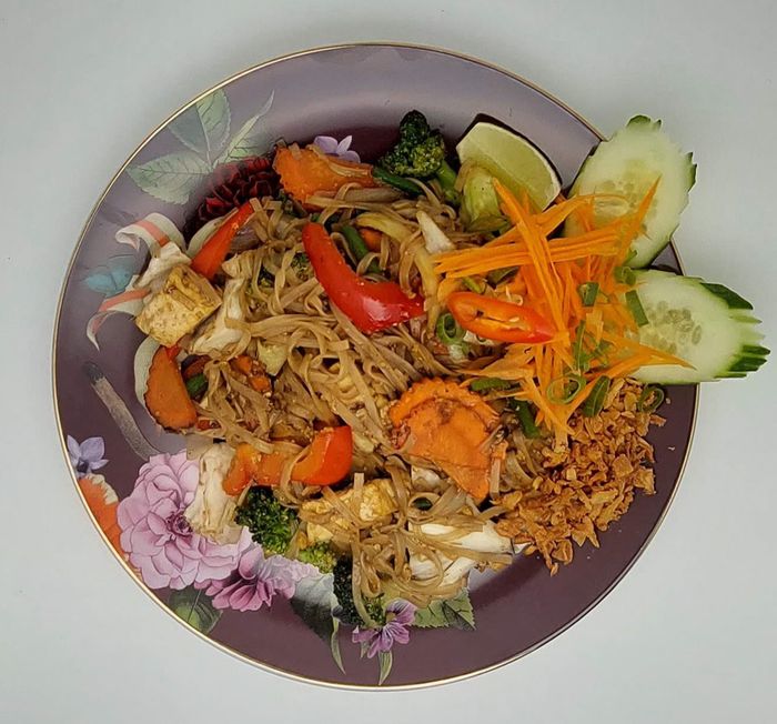 Een bord pad thai met tofu, broccoli en wortelen, gegarneerd met verse komkommer, limoen en geraspte wortel.