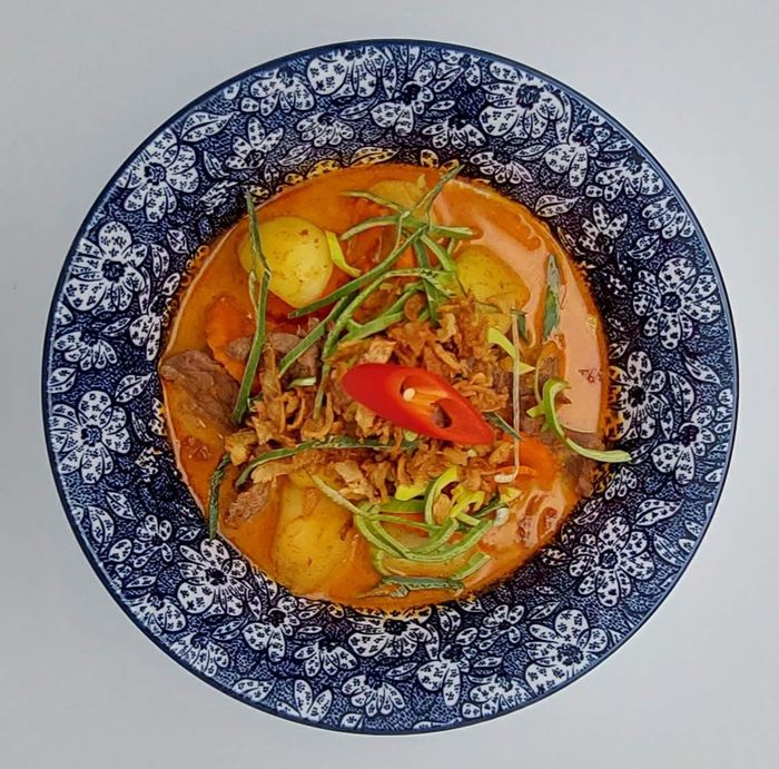 Een kom rode curry, gegarneerd met gesneden chilipeper, kruiden en krokante sjalotjes, geserveerd in een blauw-wit gestreepte kom.