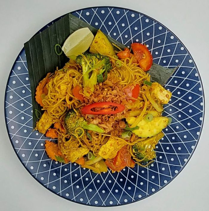 Een kom roergebakken noedels met broccoli, tofu en tomaat, gegarneerd met een schijfje chilipeper en limoen, geserveerd op een blauw bord met patroon.