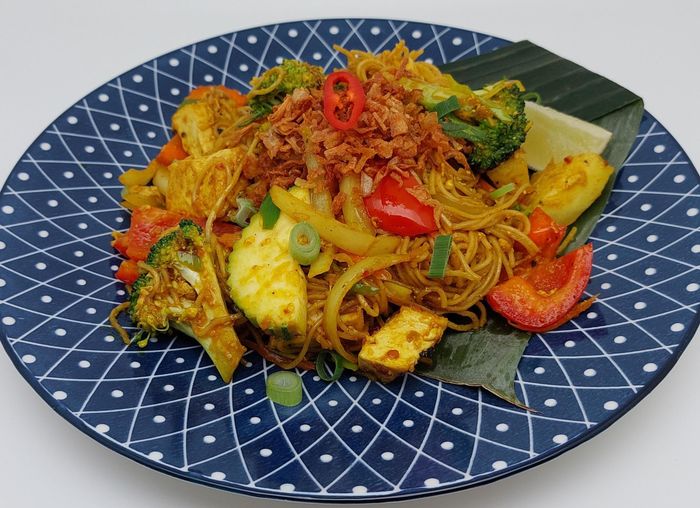 Een roerbakgerecht met noedels, groenten zoals broccoli, paprika en tofu, geserveerd op een blauw bord met een patroon en gegarneerd met een blad.