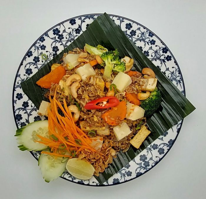 Een bord gebakken rijst met tofu, broccoli, wortels, cashewnoten en een rode chilipeper, geserveerd op een bananenblad.