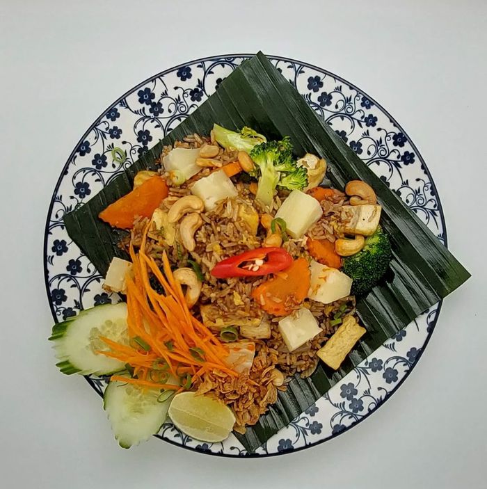 Bovenaanzicht van gebakken rijst met tofu, cashewnoten, broccoli en wortelen op een bananenblad, bovenop een blauw-wit bord met bloemenmotief.