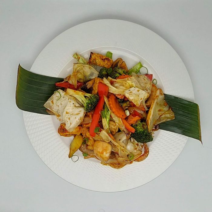 Een roerbakgerecht met tofu en groenten, geserveerd op een wit bord en gegarneerd met twee bananenbladeren.