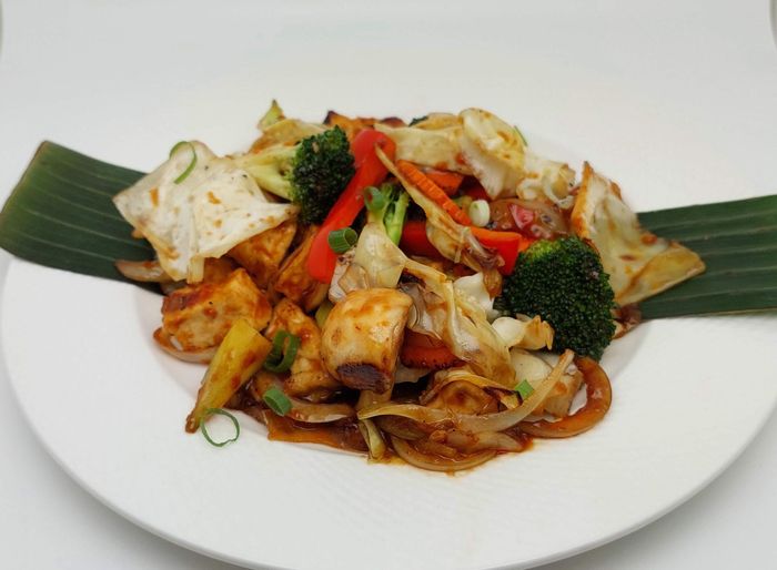 Een roerbakgerecht met tofu, broccoli, paprika en uien, geserveerd op een wit bord bekleed met bananenbladeren.