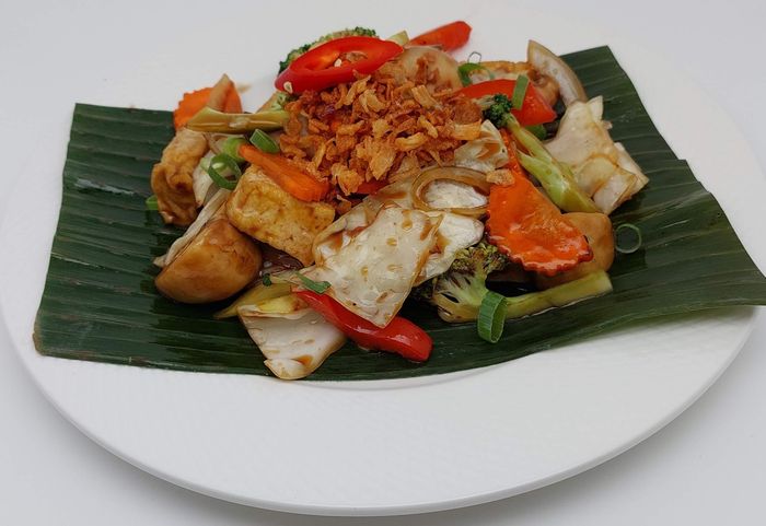 Gebakken tofu en groenten, gegarneerd met krokant gebakken sjalotjes, geserveerd op een groen bananenblad op een wit bord.
