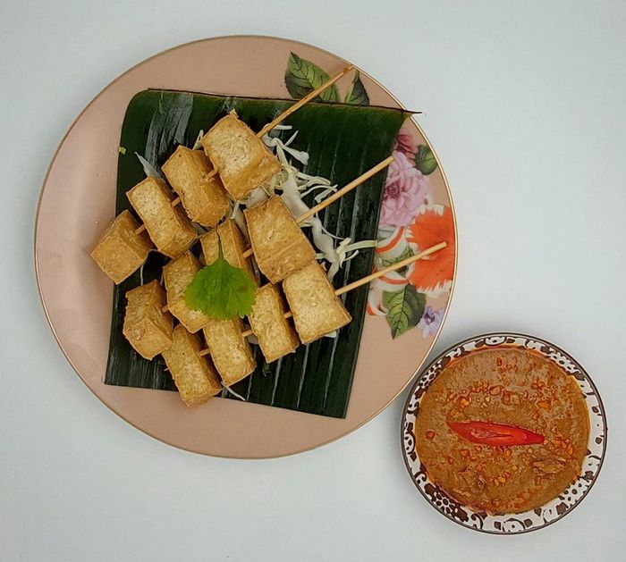 Gefrituurde tofu-spiesjes op een bananenblad, geserveerd op een bord met bloemmotief en een dipsausje van pindasaus.