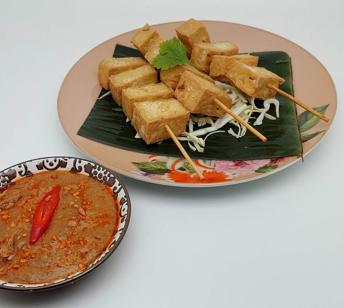 Gegrilde tofu-spiesjes op een bord bekleed met bananenblad, met een dipsausje en een chilipeper als garnering.