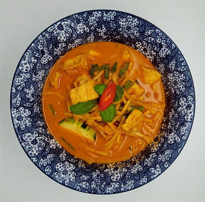 Een kom romige, oranje Thaise rode curry met kip, groenten en een garnering van rode chilipeper, geserveerd in een blauwe kom met patroon.