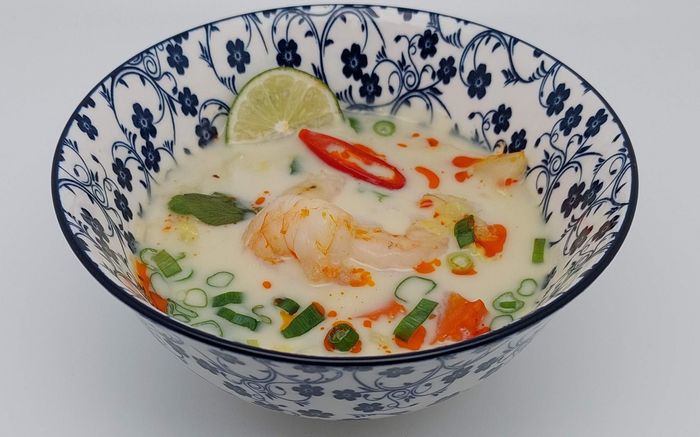 Een kom Thaise kokossoep met garnalen, een schijfje limoen, rode chilipeper en lente-uitjes in een blauw-wit gestreepte kom.