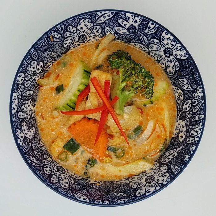 Een kom romige Thaise curry met sinaasappelsmaak, broccoli, wortels en gesneden rode chilipeper, geserveerd in een blauw-wit patroonkom.