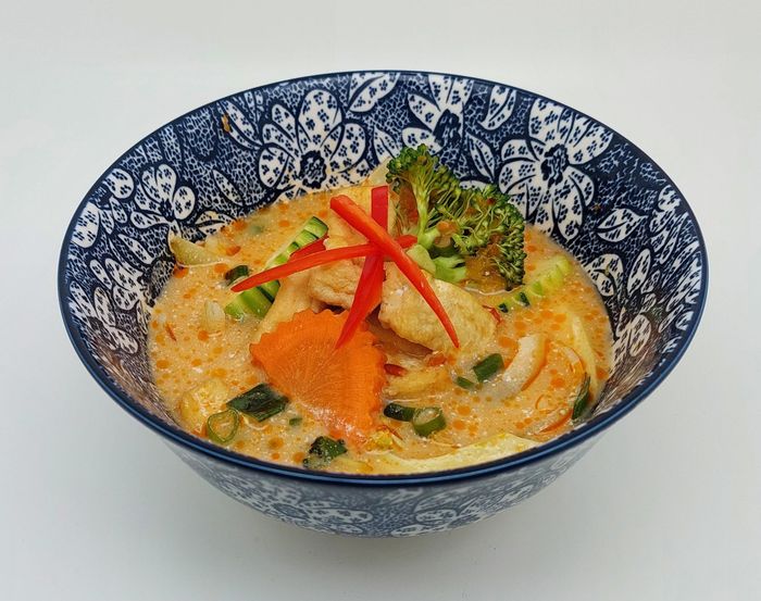 Een kom romige sinaasappelcurry met broccoli, wortels en reepjes rode paprika in een blauw-witte keramische kom met patroon.