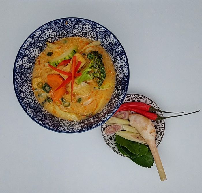 Een kom Thaise rode curry met groenten, naast een klein schaaltje met rode chilipepers, citroengras en kaffirlimoenblaadjes.
