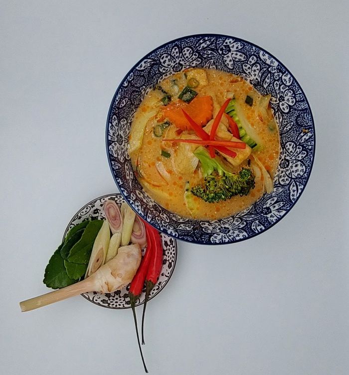 Een blauw-wit gestreepte kom Thaise curry met groenten, naast een klein schaaltje met aromatische ingrediënten zoals chilipepers en kruiden.
