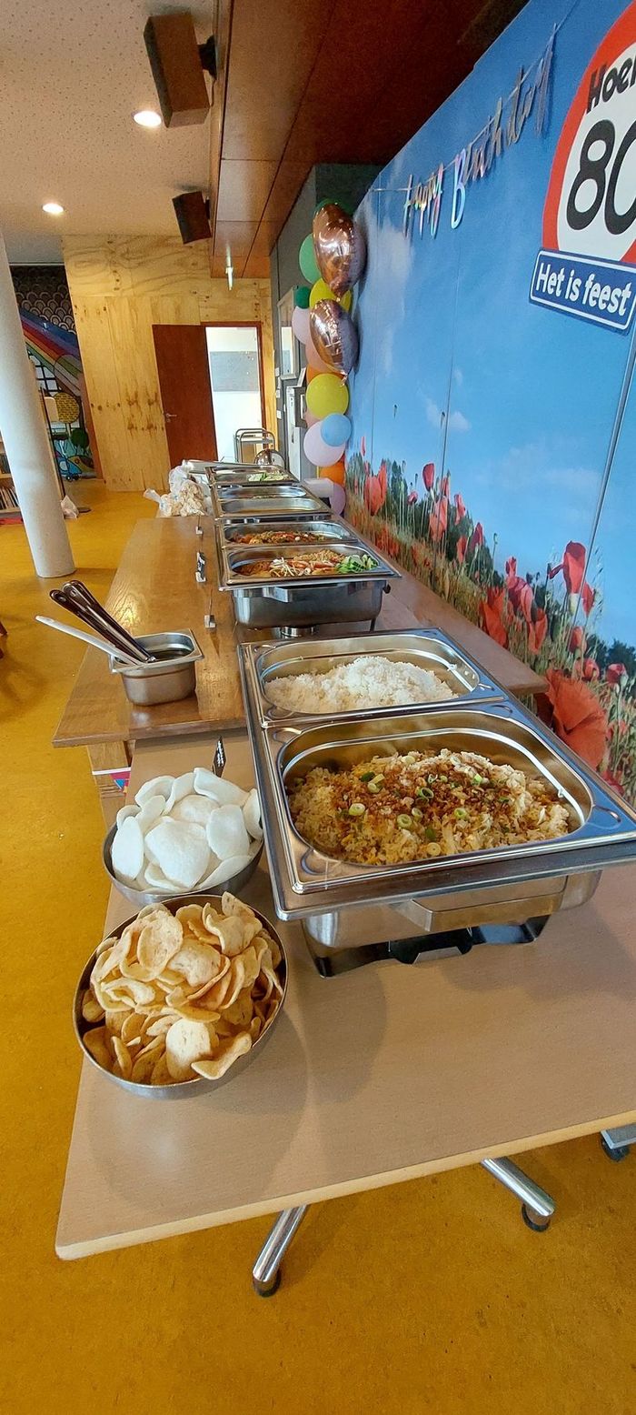 Een buffettafel vol warmhoudschalen met eten en schalen met crackers, opgesteld voor een decoratie voor een 80e verjaardagsfeest.