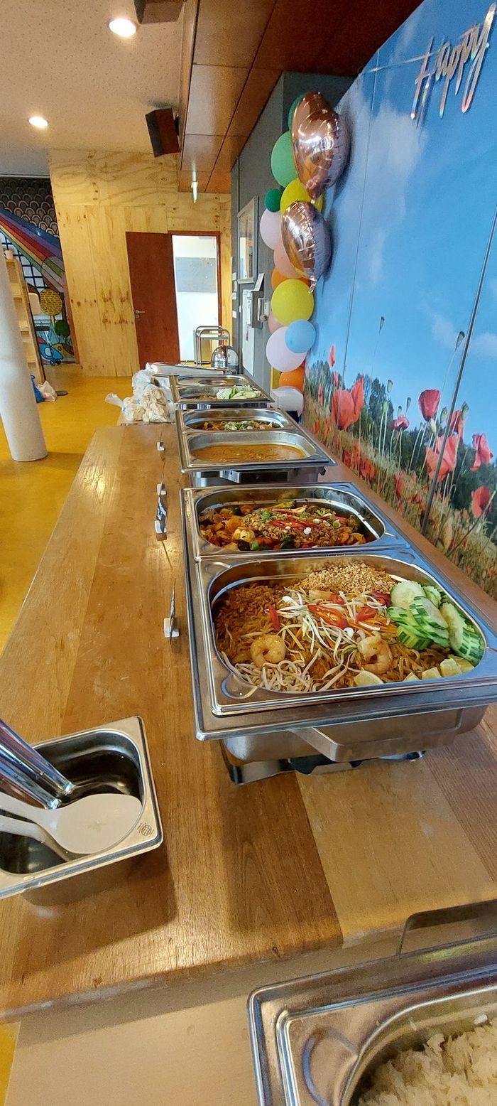 Een buffet met metalen serveerschalen vol bereid voedsel, opgesteld tegen een muur versierd met kleurrijke ballonnen.