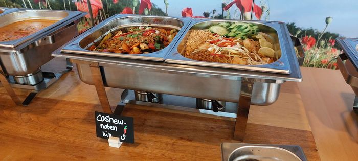 Een warmhoudschaal van roestvrij staal met twee compartimenten voor het serveren van gerechten, voorzien van een klein zwart etiket, op een houten tafel.