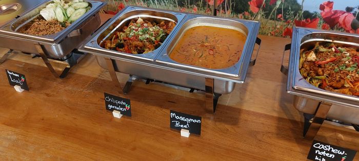 Een buffettafel met drie metalen warmhoudschalen gevuld met diverse gekookte gerechten, elk met een klein etiketje ervoor.