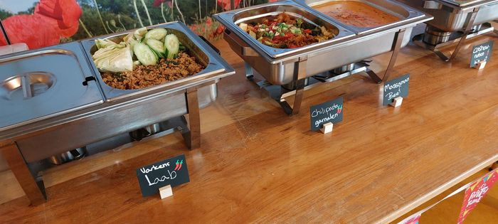 Cateringbuffet met roestvrijstalen warmhoudschalen en kleine krijtbordjes met labels op een houten tafel.