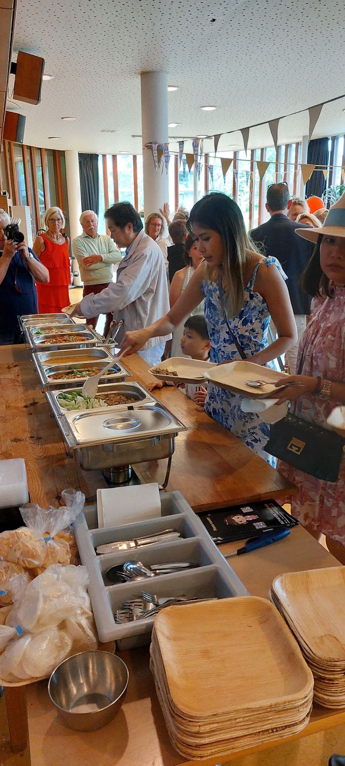 Mensen scheppen zelf hun eten op bij een buffet met roestvrijstalen warmhoudbakken in een lichte, sfeervol ingerichte evenementenruimte.