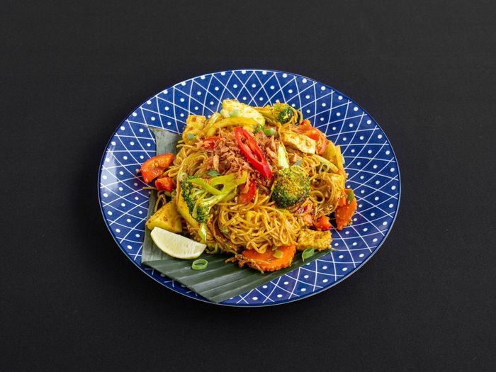 Een roerbakgerecht met noedels, broccoli, wortels en chili, geserveerd op een bananenblad in een blauw, gedessineerd keramisch bord.