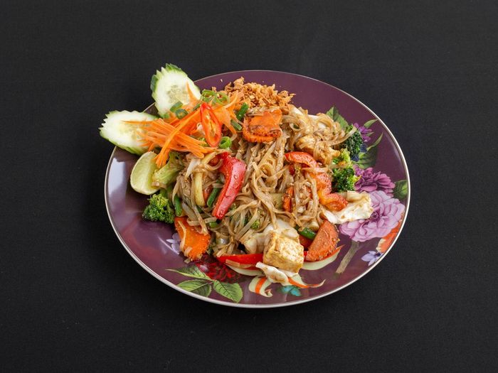 Een kleurrijk bord met roergebakken Pad Thai met tofu, groenten, chili, limoen en komkommer op een zwarte achtergrond.