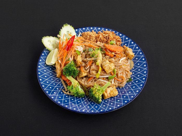 Een blauw bord met een wit geometrisch patroon, belegd met roergebakken noedels, tofu, broccoli en plakjes komkommer.