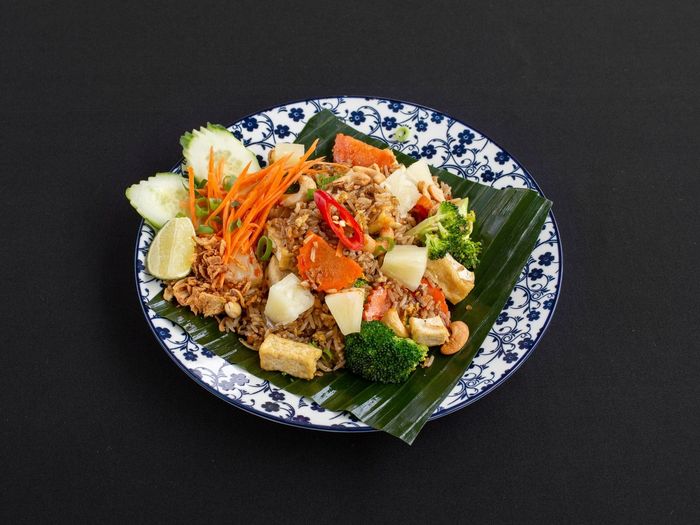 Thaise ananas-gebakken rijst met tofu, broccoli, wortelen en komkommerschijfjes, geserveerd op een bananenblad op een gedecoreerd bord.