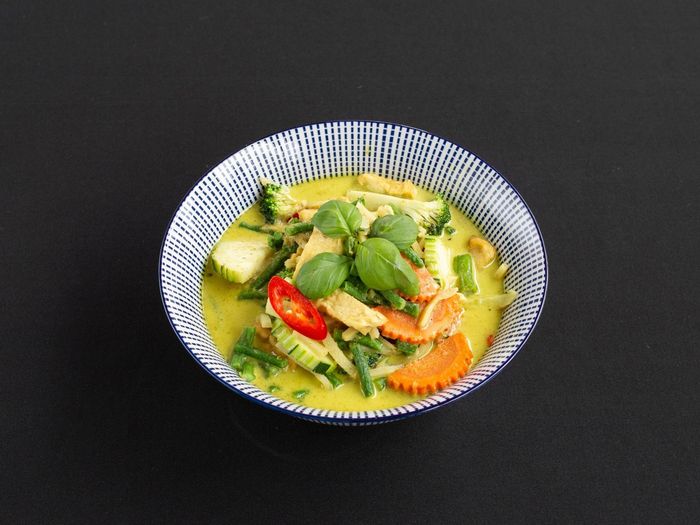 Een kom groene curry met groenten, gesneden wortelen, rode chilipeper en verse basilicumblaadjes tegen een donkere achtergrond.