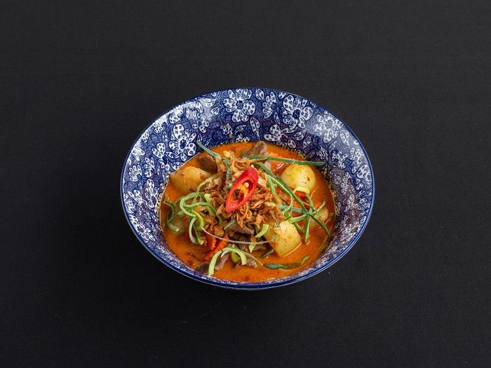 Een blauw-wit gestreepte kom met rode Thaise curry, vlees, aardappelen en gesneden rode chilipeper, op een zwarte achtergrond.