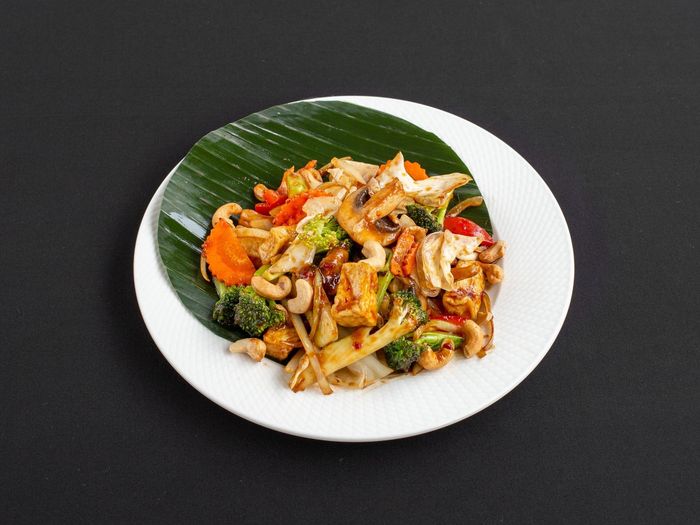 Een roerbakgerecht van groenten, tofu en cashewnoten, geserveerd op een bananenblad op een wit bord tegen een zwarte achtergrond.