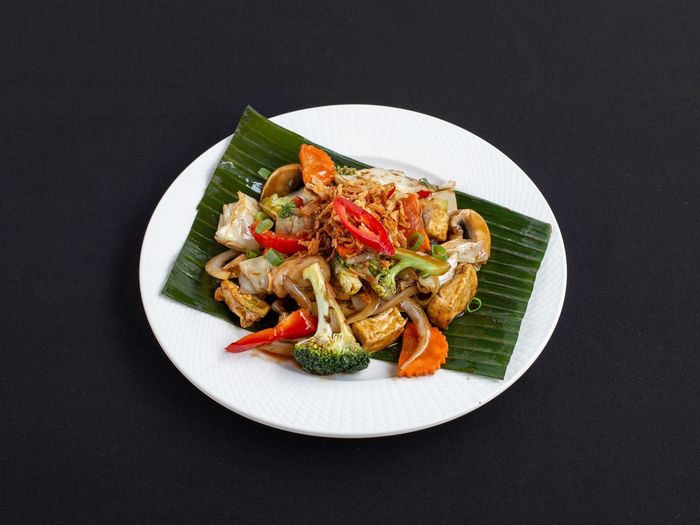 Een roerbakgerecht van groenten en tofu, geserveerd op een bananenblad op een wit bord tegen een zwarte achtergrond.