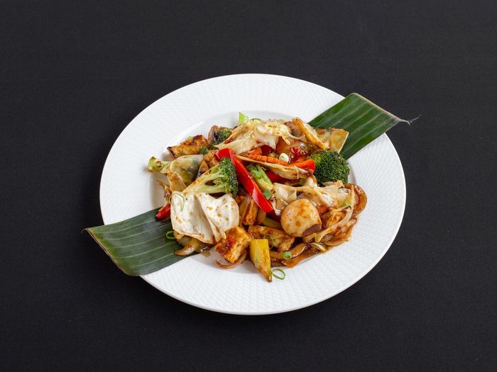 Een roerbakgerecht met broccoli, paprika en tofu, geserveerd op een bananenblad op een wit bord tegen een zwarte achtergrond.