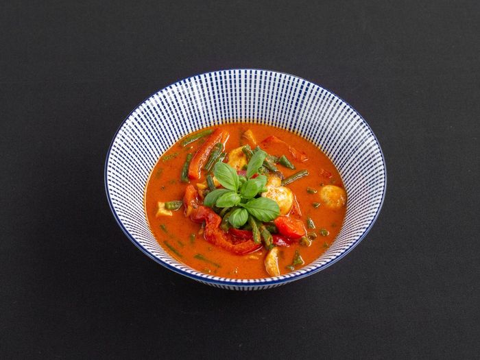 Een kom felrode curry met groenten en basilicum, geserveerd in een blauw-wit patroonkom op een zwarte ondergrond.