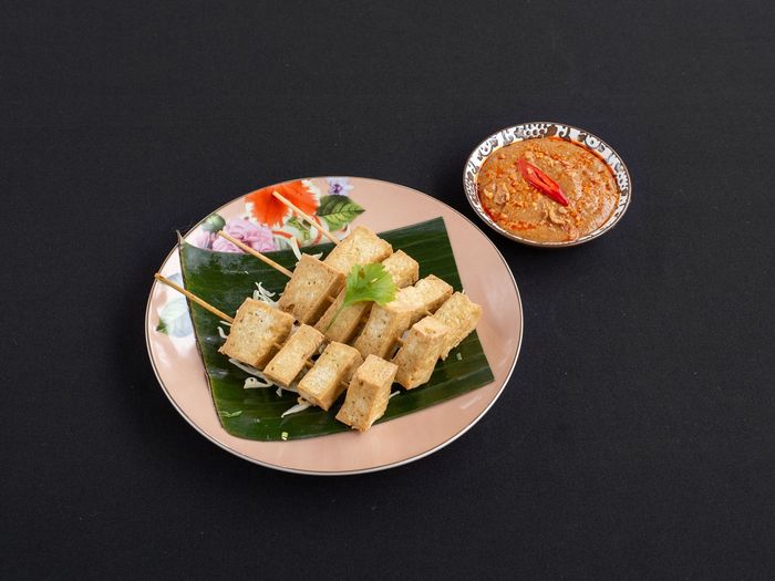 Gegrilde tofu-spiesjes op een met bananenblad bekleed bord met een dipsausje op een zwarte ondergrond.