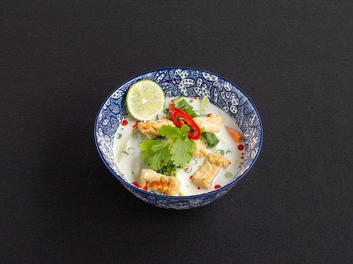 Een blauw-witte kom met patroon, gevuld met romige Tom Kha-soep, gegarneerd met tofu, koriander, limoen en plakjes rode chilipeper.