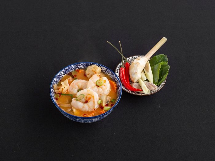 Een kom pittige garnalentomyumsoep naast een klein schaaltje met aromatische ingrediënten zoals chili, citroengras en kaffirlimoenblaadjes.