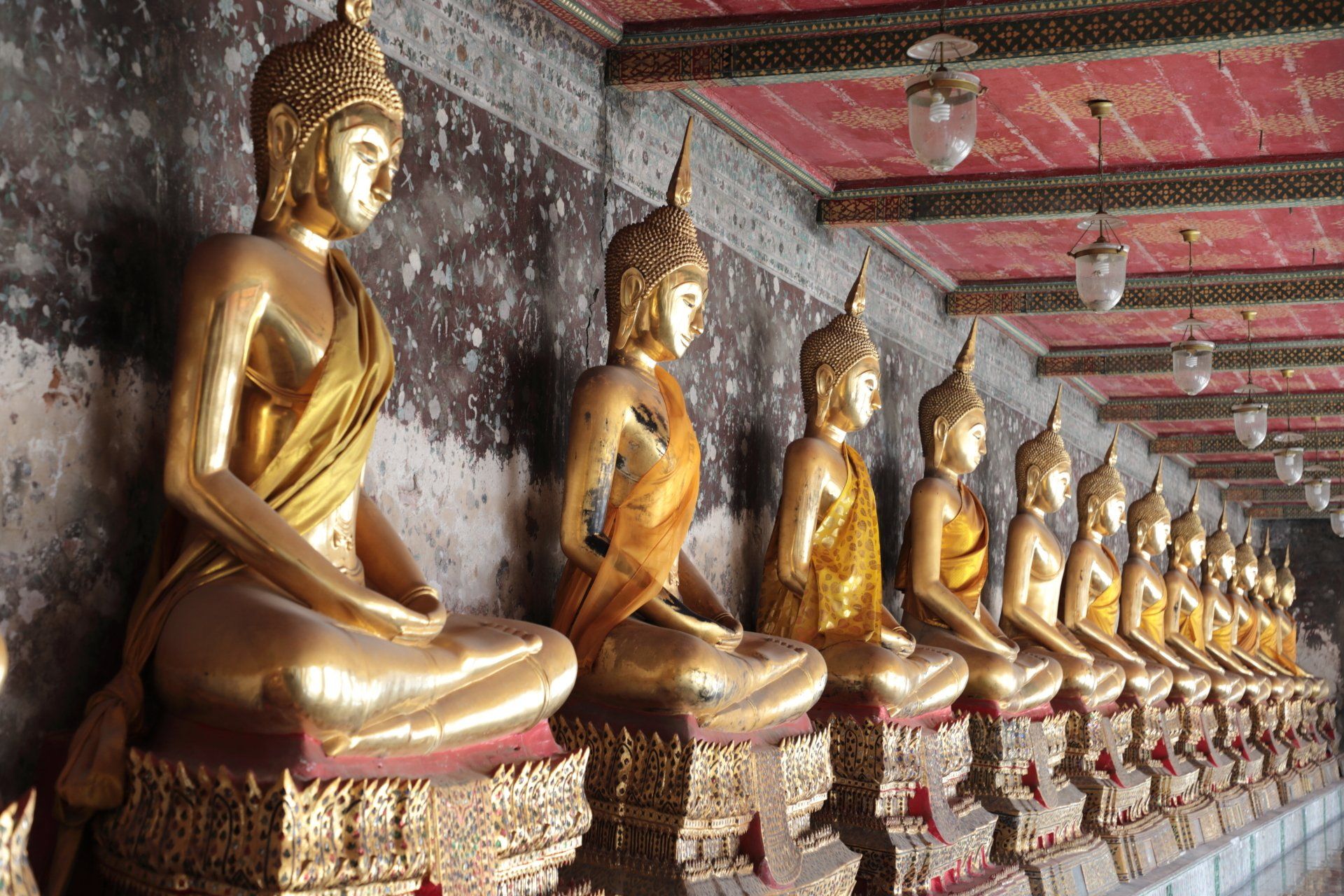 Een rij vergulde Boeddhabeelden in meditatiehoudingen staat langs een gang met een plafond met patronen en verweerde muren.