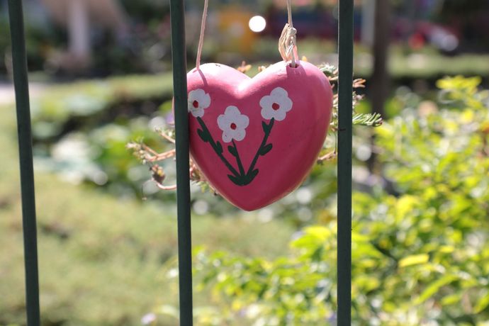 Een roze, hartvormige decoratie met witte bloemen hangt aan een metalen hek in een tuin.