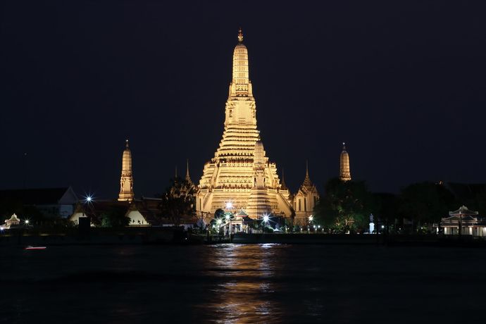De Wat Arun-tempel is 's nachts verlicht en de gouden, gloeiende torenspitsen weerspiegelen zich in het donkere water van de Chao Phraya-rivier.