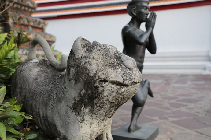 Op de voorgrond staat een verweerd stenen dierenbeeld, met op de achtergrond een donkermetalen figuur in een biddende houding.