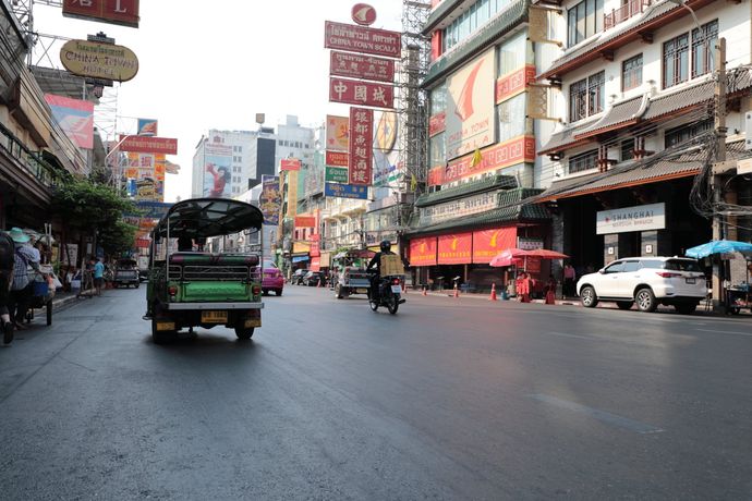 Straatbeeld in Bangkoks Chinatown met een tuk-tuk, een motorrijder en kleurrijke uithangborden onder een heldere, wazige hemel.