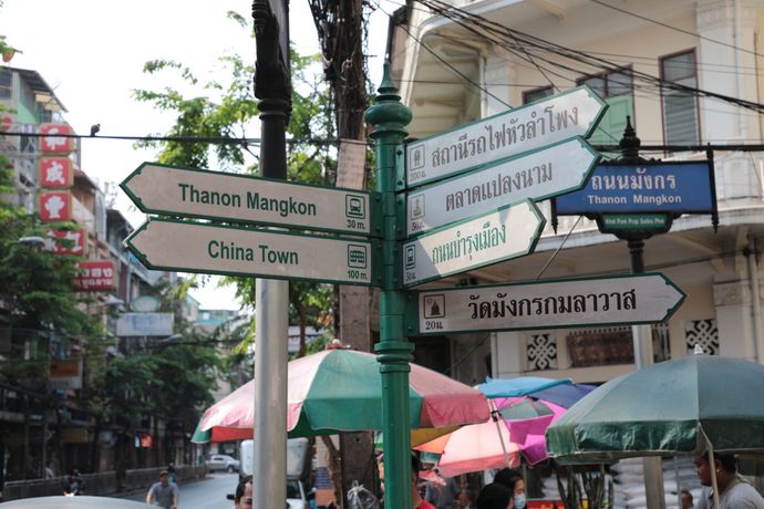 Een straatnaambord in Bangkoks Chinatown toont in het Thais en Engels de route naar nabijgelegen bezienswaardigheden, tegen de achtergrond van een drukke straat.