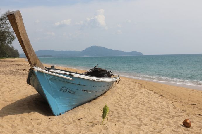 Een lichtblauwe houten vissersboot ligt op een zandstrand vlakbij de oceaan, met in de verte een berg zichtbaar.
