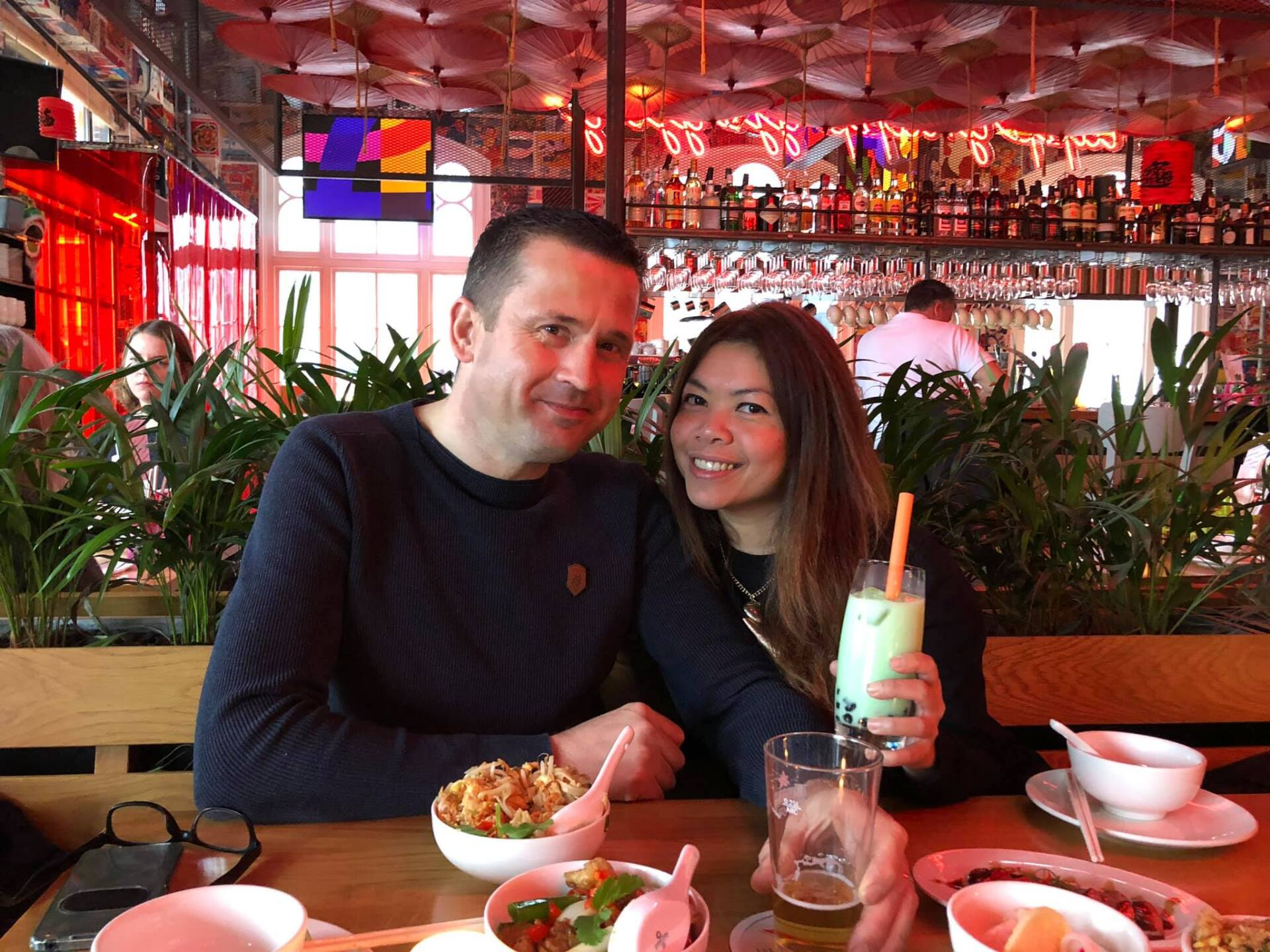 Een stel zit glimlachend aan een houten restauranttafel, met eten en drinken voor zich onder een overkapping van parasols.