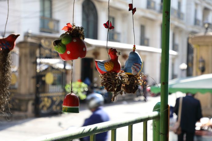 Hangende decoratieve objecten, waaronder rode en blauwe vogelfiguren, hangen voor een drukke, zonovergoten stadsstraat.